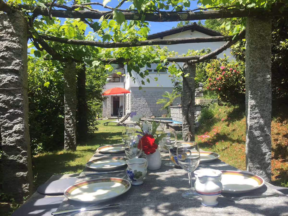 Table en pierre dressée sous la pergola avec parasol rouge, dans le jardin de la villa